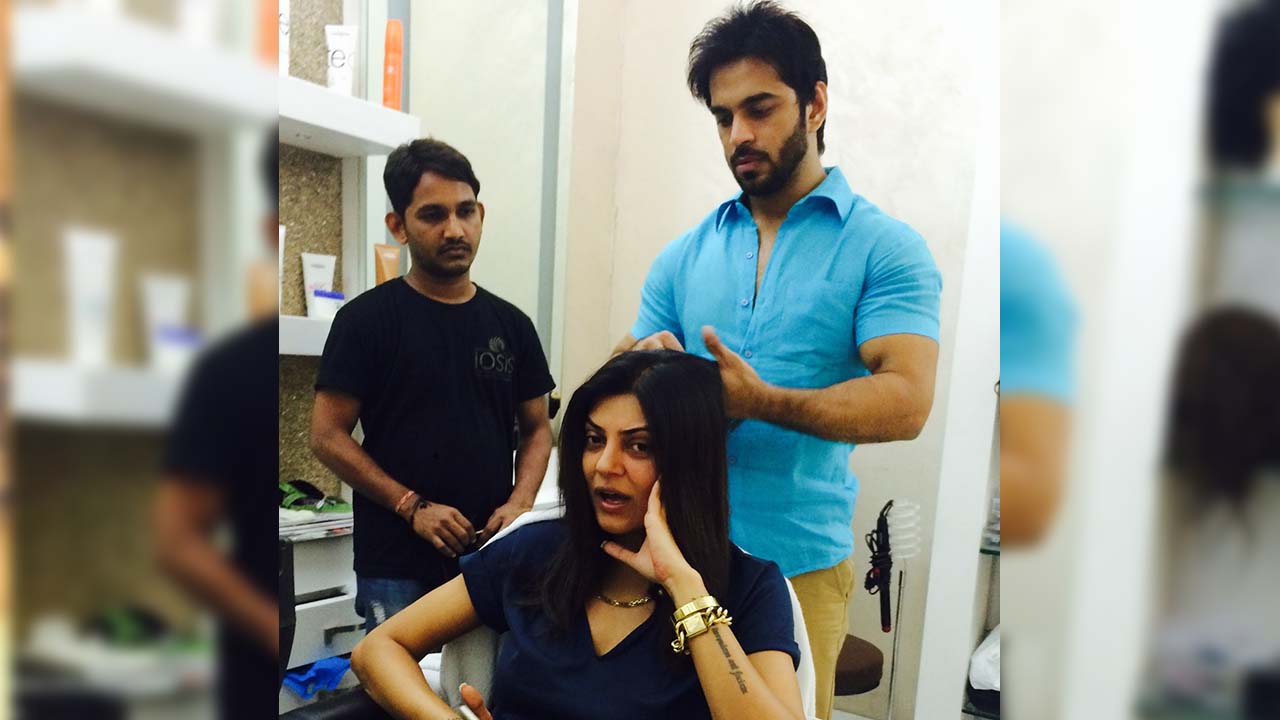 "A hairstylist in a blue shirt working on a woman's hair in a salon, while another staff member in a black t-shirt observes in the background."